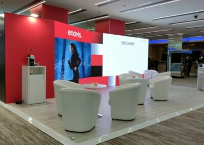 Stand ENOVIS al congresso SIOT 2024. Lo stand presenta una struttura moderna con pareti bianche e blu, loghi ENOVIS prominenti e illuminazione a LED. Tavolini e sedute bianche per incontri, materiali informativi su tavoli espositivi. L'ambiente è professionale e tecnologico, con pannelli grafici che illustrano prodotti medici e soluzioni avanzate.