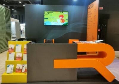 Uno stand per eventi con un design aperto e arioso, pensato per mettere in risalto i prodotti e servizi dell'espositore. È allestito con uno schermo digitale per presentazioni e materiali promozionali, circondato da piante decorative che conferiscono un tocco naturale allo spazio. Alcuni visitatori osservano il materiale esposto, creando un'atmosfera dinamica e interattiva.