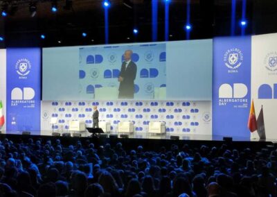 In prospettiva il palco realizzato da Key Comunicazione per conto di Federalberghi Roma in ocassione dell'Albergatore Day 2025. Spica il colore bianto tra diverse tonalità di blue sull0 sfondo del palco dove viene proiettato un video. La platea piena degli assitengi all'evento.