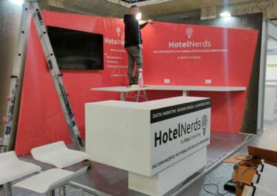 Operaio al lavoro su uno stand rosso HotelNerds con bancone bianco, attrezzi in vista e logo in evidenza. Progetto di Key Comunicazione.