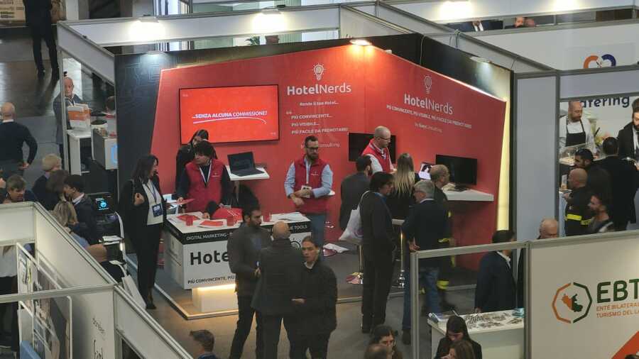 “Stand HotelNerds all’Albergatore Day 2025 con pareti divisorie in colore rosso affollato di visitatori e staff con gilet rossi durante un evento fieristico. Ideato da Key Comunicazione.