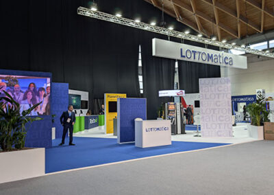 Vista generale dello stand di Lottomatica ad Enada 2025, caratterizzato da un ampio spazio con pavimento blu e pareti bianche personalizzate con il logo dell’azienda. Un grande schermo LED mostra immagini dinamiche, mentre elementi decorativi verdi aggiungono freschezza allo spazio. Il progetto è stato curato da Key Comunicazione.