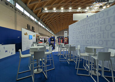 Vista interna dell’area business dello stand Lottomatica, con tavolini alti e sedie bianche, circondati da pareti con il logo ripetuto in grigio chiaro su fondo bianco. L’allestimento è stato curato nei minimi dettagli da Key Comunicazione.