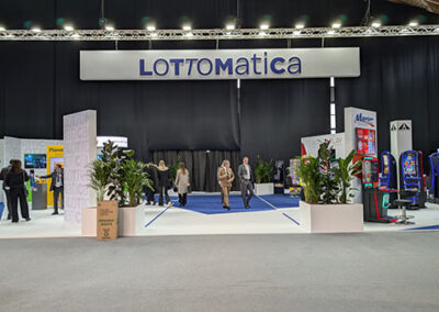 Vista generale dello stand di Lottomatica ad Enada 2025, caratterizzato da un ampio spazio con pavimento blu e pareti bianche personalizzate con il logo dell’azienda. Un grande schermo LED mostra immagini dinamiche, mentre elementi decorativi verdi aggiungono freschezza allo spazio. Il progetto è stato curato da Key Comunicazione.