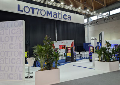 Lo stand di Lottomatica con una struttura bianca e blu, un grande logo sospeso e aree espositive con macchine da gioco. Piante decorative in vasi bianchi arricchiscono l’ambiente. L’allestimento è stato progettato da Key Comunicazione.