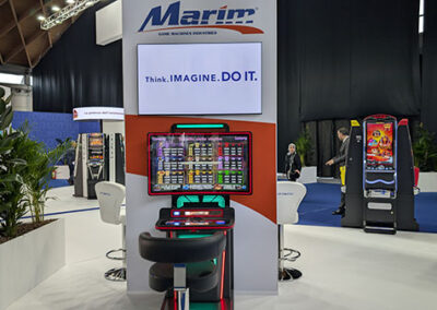 Un totem espositivo del brand Marim con un grande schermo che riporta il claim “Think. Imagine. Do It.” e una postazione interattiva con slot machine illuminate. Sullo sfondo, altre aree dello stand Lottomatica. Un progetto realizzato da Key Comunicazione.