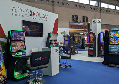 La sezione Ares Play all’interno dello stand di Lottomatica, con macchine da gioco illuminate e un grande schermo con il motto “Open your game”. Le postazioni presentano un design moderno con dettagli in rosso e nero. L’allestimento è stato curato da Key Comunicazione.