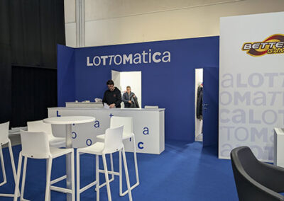 L’area reception dello stand Lottomatica, con un ampio bancone bianco su sfondo blu. Tavoli alti bianchi e sgabelli coordinati offrono uno spazio per incontri di lavoro. Il design è stato sviluppato da Key Comunicazione.