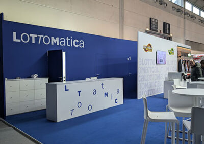 Un’altra angolazione della reception dello stand Lottomatica, con una parete blu personalizzata e un desk accoglienza. Lo spazio presenta un’illuminazione strategica per valorizzare il brand. Progetto curato da Key Comunicazione.