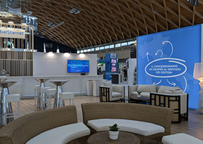 Vista panoramica dello stand di Global Starnet ad Enada 2025, con zona lounge, area bar e ampi spazi espositivi. Il grande pannello blu con grafiche direzionali enfatizza la comunicazione del brand, mentre le sedute in rattan e gli sgabelli bianchi aggiungono un tocco elegante. Il design è stato curato da Key Comunicazione.