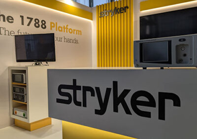 Diverso punto di vista dello stand Stryker con colori predominanti bianco e giallo. Il logo "Stryker" è ben visibile su una parete laterale con pannelli verticali gialli. Sullo sfondo, il testo "The 1788 Platform – The future in your hands" è evidenziato in nero e giallo. Davanti, un bancone con il logo dell’azienda e una piccola area con TV e dispositivi tecnologici per la presentazione dei prodotti. La Key Comunicazione ha curato l’allestimento