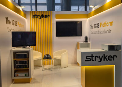 Altro anglolo dello s tand espositivo Stryker con colori predominanti bianco e giallo. Il logo "Stryker" è ben visibile su una parete laterale con pannelli verticali gialli. Sullo sfondo, il testo "The 1788 Platform – The future in your hands" è evidenziato in nero e giallo. Davanti, un bancone con il logo dell’azienda e una piccola area con TV e dispositivi tecnologici per la presentazione dei prodotti. La Key Comunicazione ha curato l’allestimento