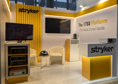 Stand espositivo Stryker con colori predominanti bianco e giallo. Il logo "Stryker" è ben visibile su una parete laterale con pannelli verticali gialli. Sullo sfondo, il testo "The 1788 Platform – The future in your hands" è evidenziato in nero e giallo. Davanti, un bancone con il logo dell’azienda e una piccola area con TV e dispositivi tecnologici per la presentazione dei prodotti. La Key Comunicazione ha curato l’allestimento.