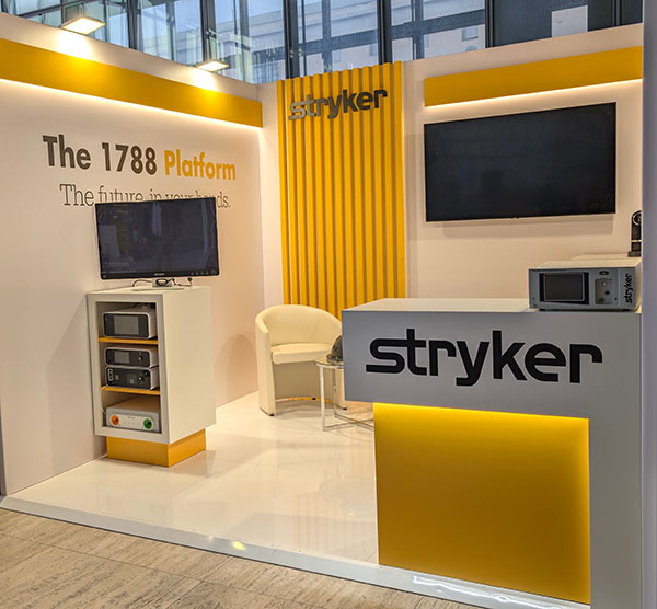 Primo piano dello Stand Stryker all ESGO 2025, con focus sul bancone bianco con dettagli gialli e il logo aziendale in nero. Sopra il bancone è posizionato un dispositivo medico. Sul lato sinistro, un mobile espositivo bianco con vari strumenti e una TV montata a parete. La Key Comunicazione ha realizzato il design dell’evento.